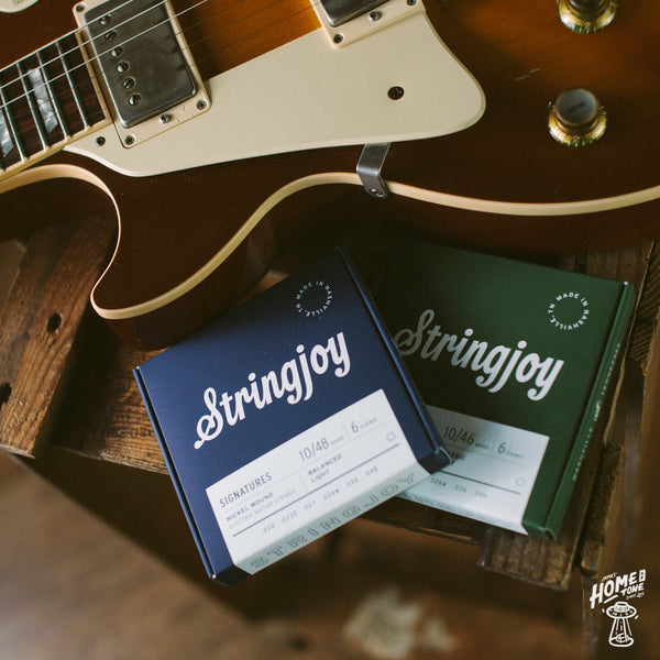 A flat lay style photograph featuring 'Stringjoy' brand guitar string boxes aside a Gibson Les Paul sunburst standard