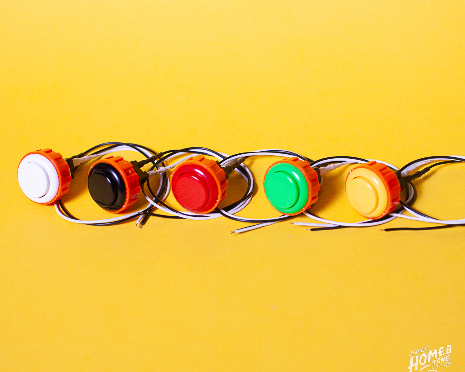 A series of colored Tesi Switch arcade buttons arranged in a line on a yellow background, each button connected to a wire.