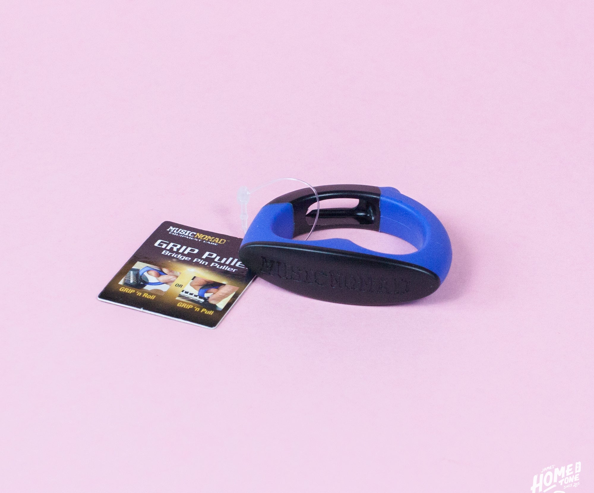 A MusicNomad GRIP Puller, a bridge pin puller tool with a blue handle, lying on a pink surface with its packaging tag visible to the left.
