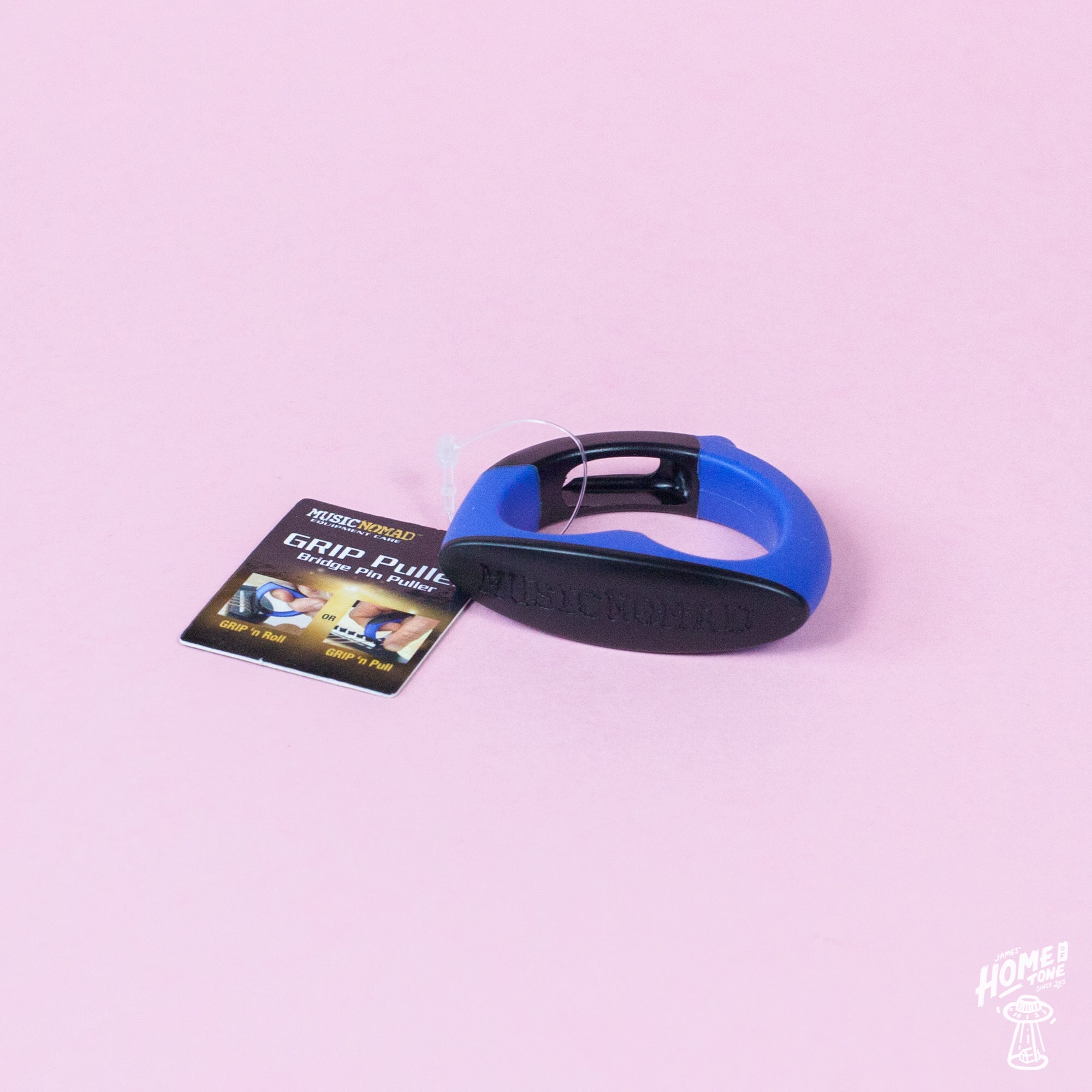 A MusicNomad GRIP Puller, a bridge pin puller tool with a blue handle, lying on a pink surface with its packaging tag visible to the left.