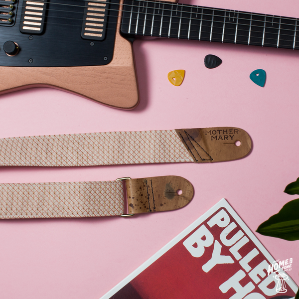 Flat lay photograph of a 'Mother mary Company' brand guitar strap, on a pink background with a boutique guitar, plectrums and a vinyl album lay out next to it.