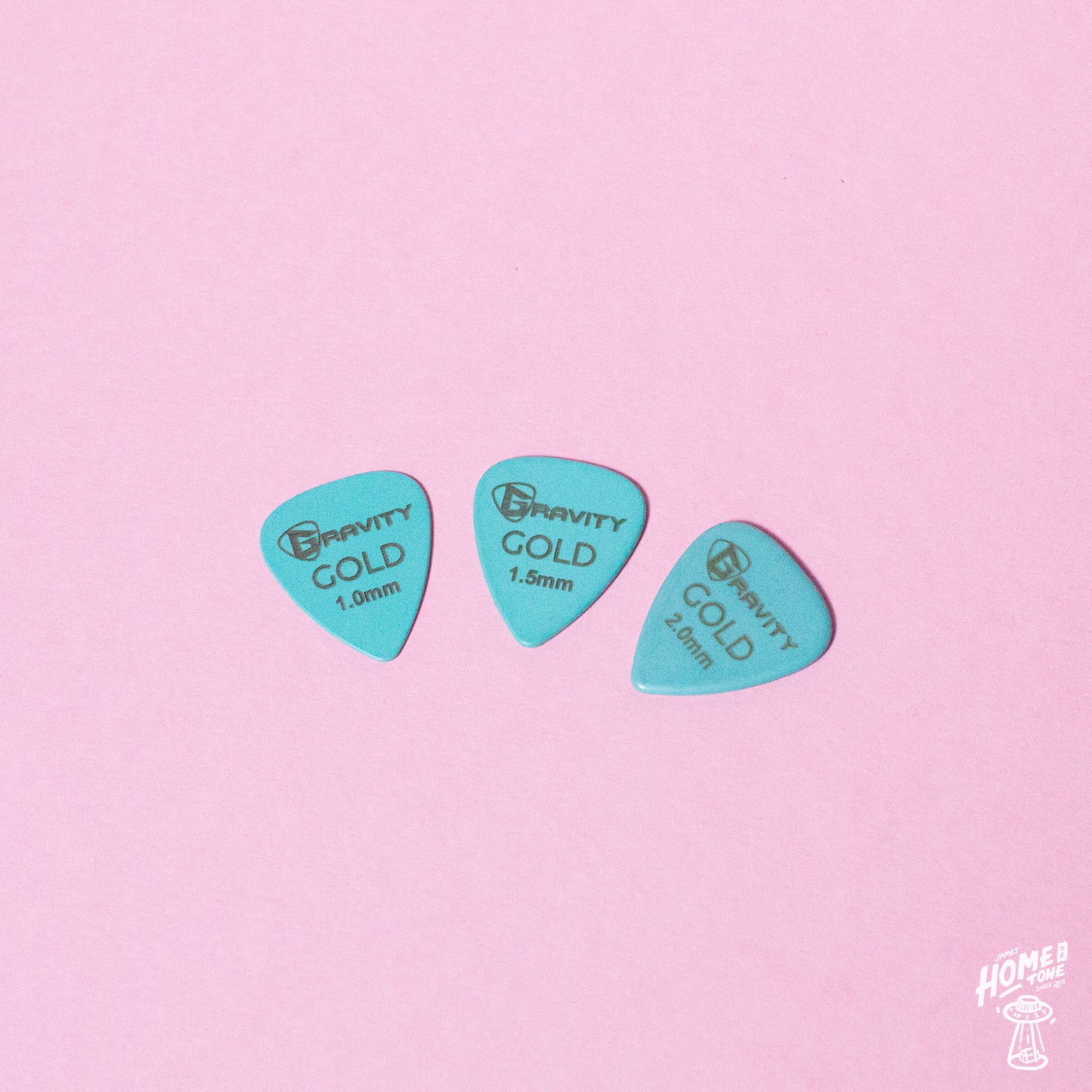 Three teal-colored guitar picks from the Gravity Gold series placed on a pink background.