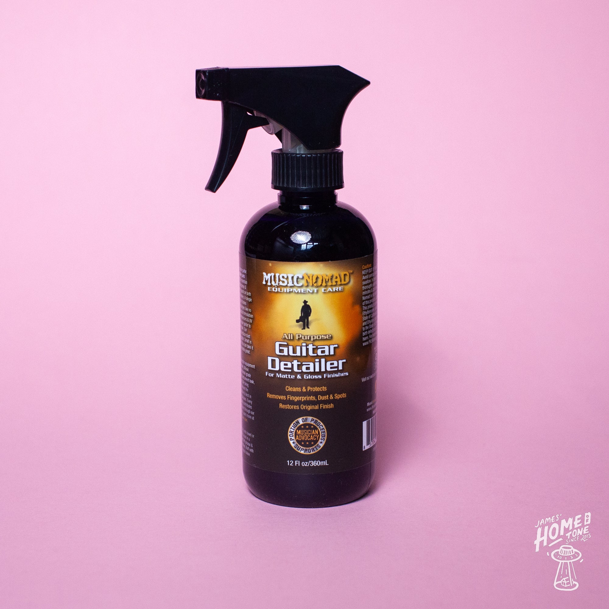 A black spray bottle with a label reading 'Music Nomad Guitar Detailer', designed for cleaning and maintaining matte and gloss finished instruments, displayed against a pink background.