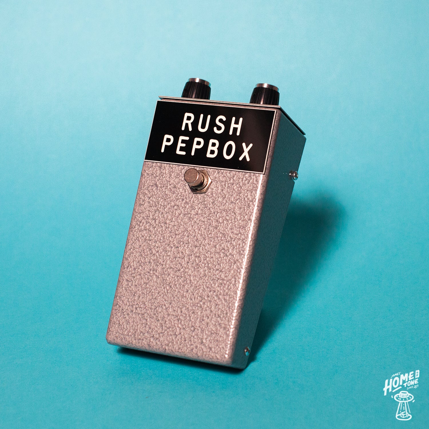 A silver Rush Pepbox fuzz pedal with black knobs and engraved panels, on a blue background.