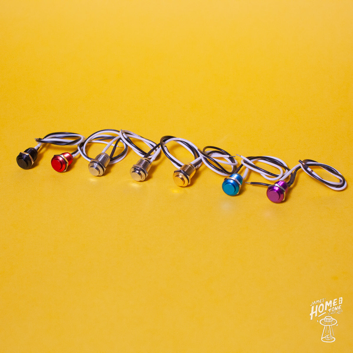 A selection of Tesi Switch momentary push button kill switches for guitars, displayed in a row on a yellow background, with each switch having a different color button and housed in metal.