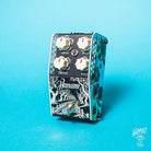 A ThorpyFX Pedals Hanami Ge Fuzz pedal with four knobs on the top for volume, boost, treble, and bass control, placed against a blue background with a white and black geometric pattern on its pearlised white base.