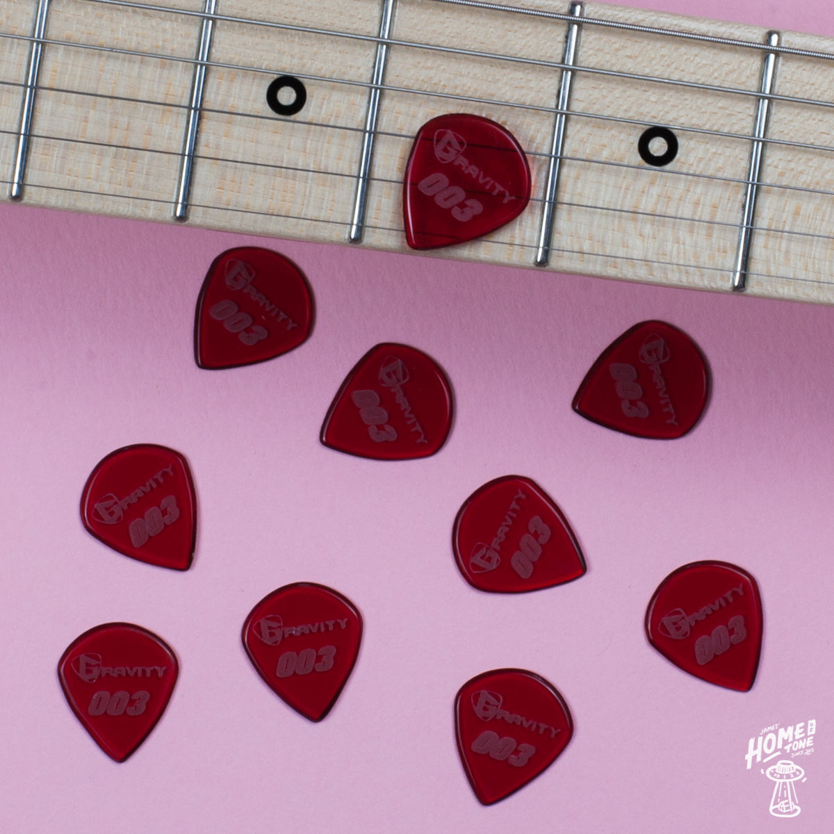 A collection of red, hand-shaped guitar picks from Gravity's Signature Series placed on a pink surface with a guitar in the background.