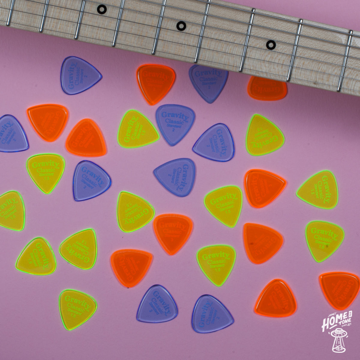 An array of colorful acrylic Gravity Classic Standard guitar picks in various shades of red, orange, yellow, and blue, laid out on a pink surface next to a guitar fretboard.