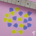 A collection of yellow and blue acrylic Gravity guitar Sunrise picks laid out on a pink surface next to a guitar neck.
