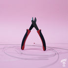 A pair of GrooveTech String Cutters with black handles and red accents, positioned above guitar strings against a pink background.