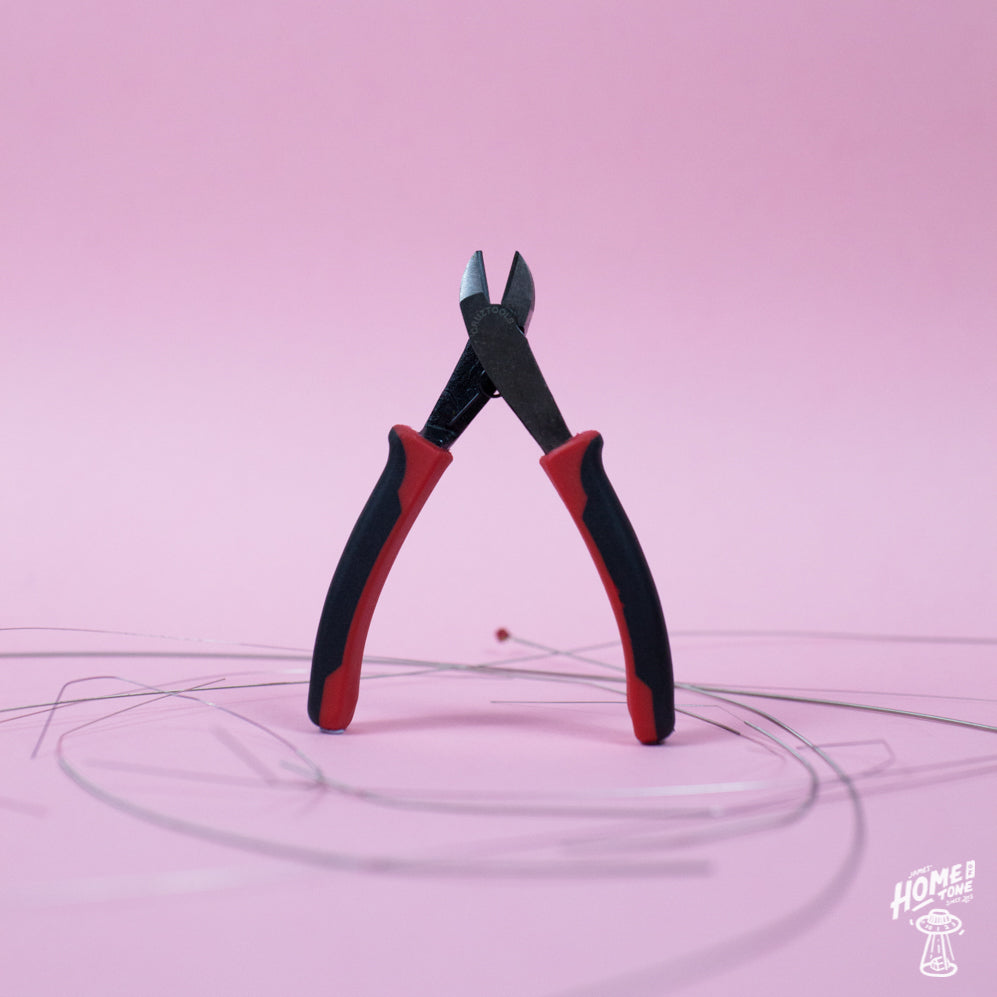 A pair of GrooveTech String Cutters with black handles and red accents, positioned above guitar strings against a pink background.